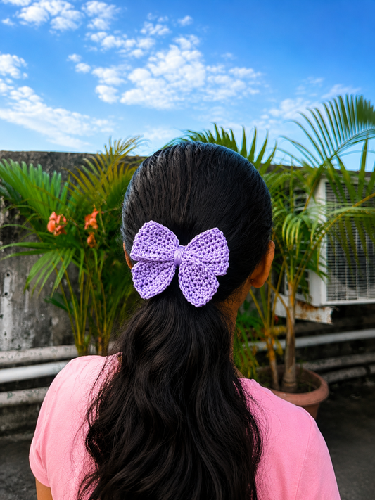 Handmade lavender crochet bow hair clip attached to a ponytail with a garden backdrop.