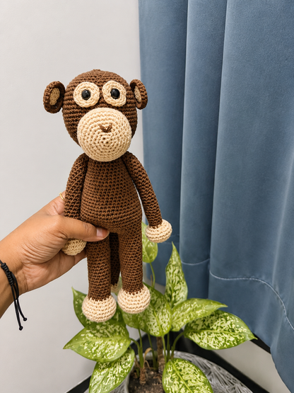 Hand holding a handmade brown crochet monkey plush toy with beige face, hands, and feet, photographed indoors beside blue curtains and green houseplants.