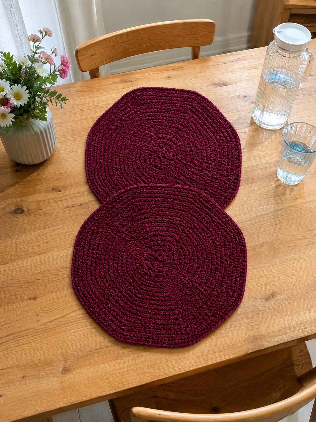 Set of two handcrafted burgundy crochet table mats made from soft velvet chenille yarn displayed on a light background with decorative greenery