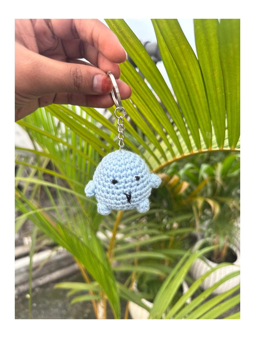 Hand holding a blue crochet keychain with a face against a green leafy background