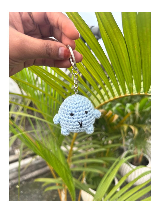 Hand holding a blue crochet keychain with a face against a green leafy background