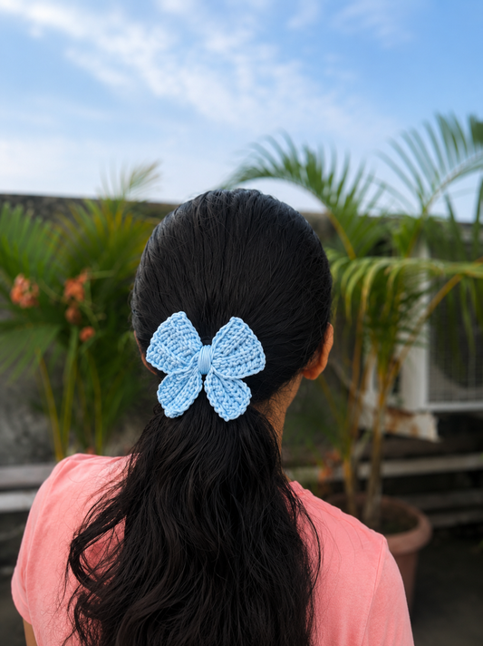 Handmade sky blue crochet bow hair clip styled on a ponytail with a natural outdoor background.