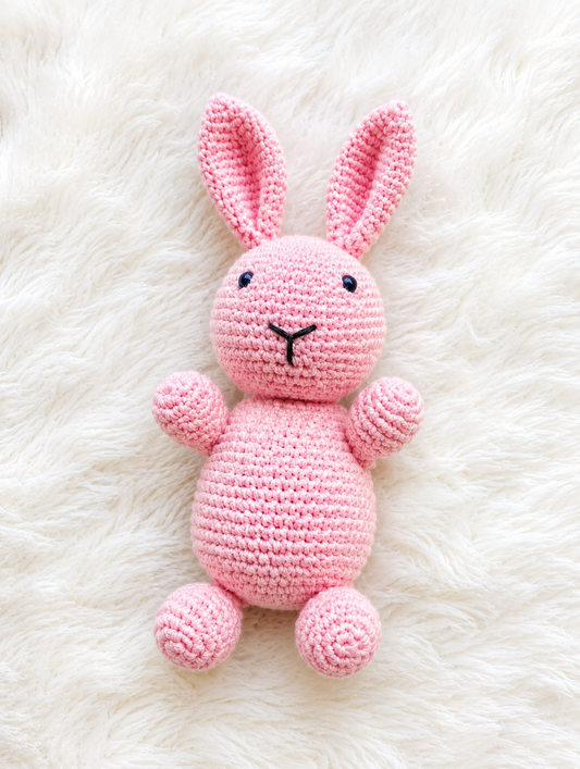 Handmade pink crochet bunny plush toy with long ears lying on a soft white furry background.