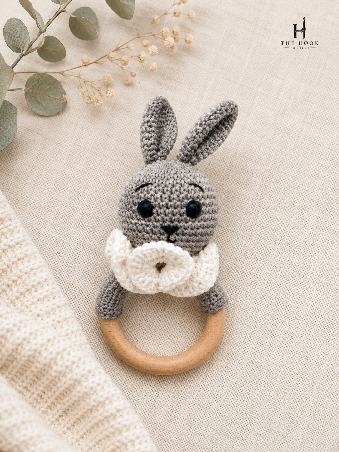 Handmade grey crochet bunny teether rattle with natural wooden ring on a neutral fabric background.